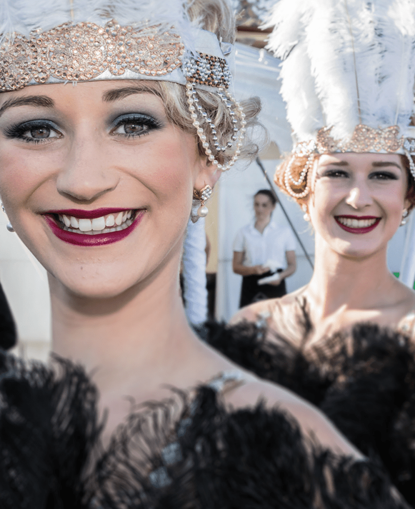 A woman dressed in a black feather dress with a white feather headdress smiles with red lipstick on